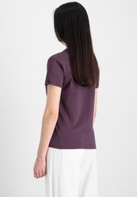 T-shirt violet à manches courtes avec un col rond, en tissu de coton lisse, et une petite étiquette logo sur la manche gauche. Pantalon blanc partiellement visible.