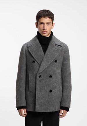 Junger Mann trägt einen grauen doppelt geknöpften Wollmantel über einem schwarzen Rollkragenpullover vor einem schlichten weißen Hintergrund.