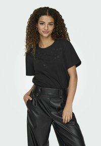 Camiseta negra de manga corta con un diseño de corte floral en el pecho, combinada con pantalones de piel sintética negra de cintura alta.