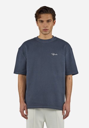 Junger Mann mit kurzen dunklen Haaren, der ein strukturiertes, dunkelblaues, übergroßes T-Shirt und eine weiße Hose trägt, und vor einem einfarbigen Hintergrund steht.