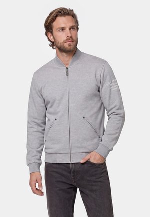 Man with beard wearing light grey zip-up jacket with pockets, standing with one hand in pocket, looking to the side.