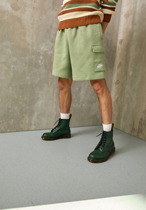 Personne portant un short cargo Nike vert clair, des chaussettes blanches, des bottes à lacets vertes et un pullover rayé, se tenant sur un sol gris.