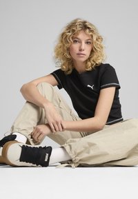 Črna kratka majica s teksturo in belim logotipom Puma, v kombinaciji s širokimi bež hlačami in črnimi supergami z belimi poudarki.