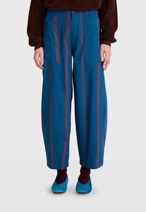 Pantalon bleu à jambes larges avec des rayures rouges verticales, porté avec des chaussettes foncées et des chaussons bleus, associé à un haut marron à manches longues.