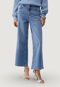 Hele sinised laiade säärtega teksapüksid kõrge vöökohaga, klassikalise viie taskuga ja peene pleekimisega. Sobitatud valgete rihmikandadega kontsadega.