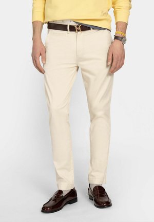 Hombre vistiendo pantalones chinos crema, cinturón de cuero marrón con hebilla dorada, sudadera amarilla, mocasines de cuero marrón, reloj de pulsera y pulseras, de pie.