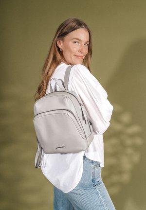 Frau mit langen Haaren, die eine weiße Bluse und blaue Jeans trägt und einen hellgrauen Rucksack über einer Schulter vor einem olivgrünen Hintergrund trägt.