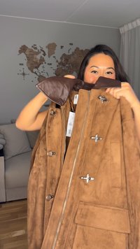 Smeđa vest od brušene kože s patentnim zatvaračem na prednjoj strani, smeđim kožnim ovratnikom i srebrnim metalnim detaljima. Ima četiri ukrasna gumba i bočni džep.