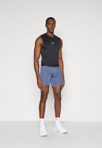 Μαύρο αμάνικο κορμάκι και μπλε αθλητικές βερμούδες. Περιέχει το λογότυπο της Nike στο κορμάκι. Φοράει λευκά αθλητικά παπούτσια και κάλτσες. Απλό φόντο.