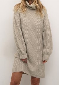 Femme portant une robe pull col roulé beige en tricot torsadé à manches longues, debout devant un fond blanc uni.