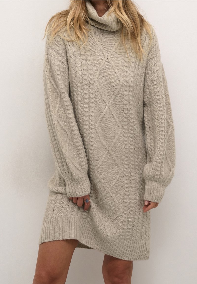 Femme portant une robe pull col roulé beige en tricot torsadé à manches longues, debout devant un fond blanc uni.