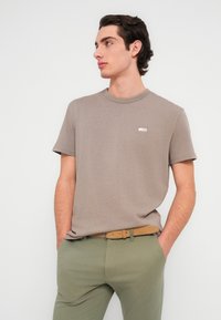 Ανοιχτό καφέ κοντομάνικο T-shirt με στρογγυλή λαιμόκοψη και μικρή λεπτομέρεια λογότυπου στο αριστερό στήθος. Συνδυάζεται με ανοιχτό πράσινο παντελόνι και ζώνη ταμπά.