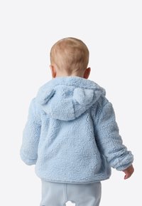 Hanorac din fleece albastru deschis, cu o suprafață moale și texturată și accente în formă de urechi pe glugă, văzut din spate, purtat de un copil.