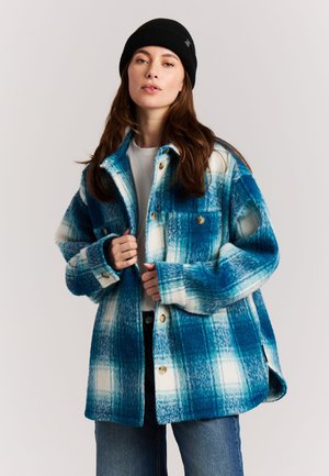 Vrouw draagt een zwarte muts, een blauw-wit geruite blouse met knopen, een wit shirt en een blauwe spijkerbroek tegen een effen achtergrond.