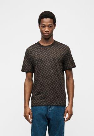 Mladý muž v tmavej vzorovanej košeli s krátkymi rukávmi a modrých džínsoch stojaci pred jednoduchým svetlým pozadím.