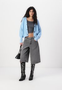 Lichtblauwe gebreide cardigan, grijs cropped top, grijze wijde shorts, zwarte kniehoge laarzen met witte borduring, met een zwarte handtas.