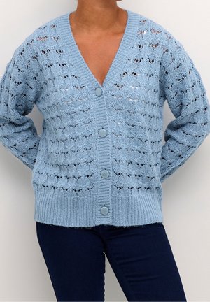 Personne portant un cardigan en maille bleu clair avec des boutons et des trous décoratifs, associé à un pantalon bleu foncé, mains dans le dos.