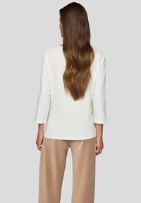 Frau mit langen, welligen braunen Haaren steht mit dem Rücken zur Kamera und trägt ein weißes Langarmshirt sowie beige Cord-Hosen mit weitem Bein vor einem grauen Hintergrund.