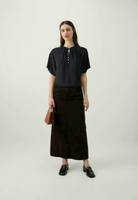 Marineblauwe blouse met korte mouwen, striknek en knopen, gecombineerd met een maxi-rok van donkerdenim. Het model houdt ook een bruine handtas vast en draagt zwarte instappers.