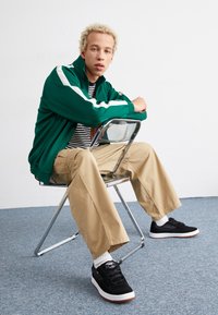 Green zip-up jacket with white stripes, striped shirt, beige wide-leg pants, and black sneakers with white soles. Sitting on a clear chair.