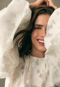 Blouse en dentelle blanche avec de grands motifs floraux texturés, dotée de manches larges et d'une coupe décontractée. Le tissu doux et transparent complète le design.