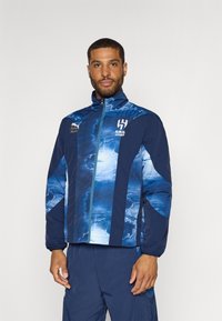 Rompevientos navy con patrón de nubes azules, cierre frontal, cuello alto, puños elásticos y logotipo bordado "AL HILAL" en el pecho.