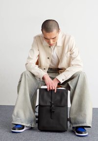 Zaino nero con chiusura a patta, realizzato in materiale liscio. Utente seduto, con giacca beige e jeans larghi chiari, su tappeto grigio.