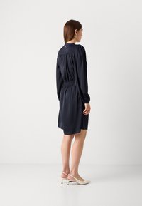 Navy blue dress with long sleeves and elastic waist, featuring a gathered back and button detail at the neckline. Paired with nude heels.