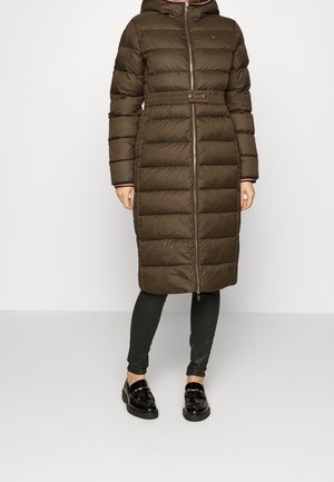 Long manteau matelassé vert olive avec ceinture à la taille et capuche, porté avec des leggings noirs et des mocassins noirs brillants.