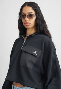 Νεαρή γυναίκα με μακριά σκούρα μαλλιά, φορά γυαλιά ηλίου, ένα μαύρο κοντό φούτερ με φερμουάρ και μπροστινές τσέπες, και ανοιχτούς μπλε τζιν.