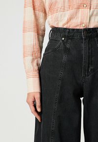 Vaqueros negros de talle alto con costuras laterales, combinados con una camisa ligera a cuadros con líneas naranjas. Detalles de botones y bolsillos visibles.