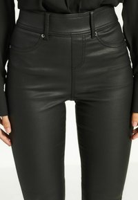 Zwarte faux leren high-waisted broek met een gladde textuur, voorzakken en zichtbare stikdetails. Strak aansluitende pasvorm.