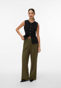 Vrouw staat, draagt een mouwloze zwarte top met knopen, olijfgroene wijde broek met riem en zwarte puntige schoenen, op een witte achtergrond.