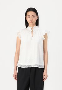 Hvit langermet bluse med volanger på skuldrene, nøkkelhull-hals, og teksturerte polka-prikker. Lett, transparent stoff med løs passform.