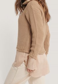 Beige Strickpullover mit strukturiertem Muster, lockerer Passform und gerippten Bündchen, über einer hellbeigen glatten Bluse getragen.