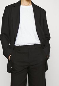 Zwarte blazer met een getailleerde pasvorm, lange mouwen en revers, gedragen over een wit T-shirt, gecombineerd met zwarte broeken. Soepele stofstructuur.