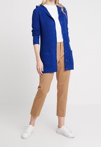 Niebieski prążkowany sweter z kapturem, zapinany na guziki z przodu i dwiema kieszeniami, zestawiony z beżowymi spodniami i białymi trampkami.