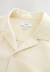Chemise crème de taille moyenne avec col, boutons sur le devant et une étiquette cousue indiquant « NEXT » et « Made in Bangladesh ».