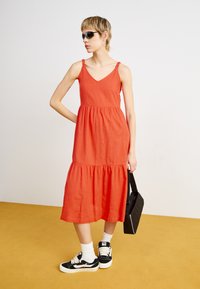 Robe maxi en lin rouge avec un décolleté en V, des bretelles ajustables et une jupe à volants ; associée à des baskets noires et un sac à bandoulière noir.