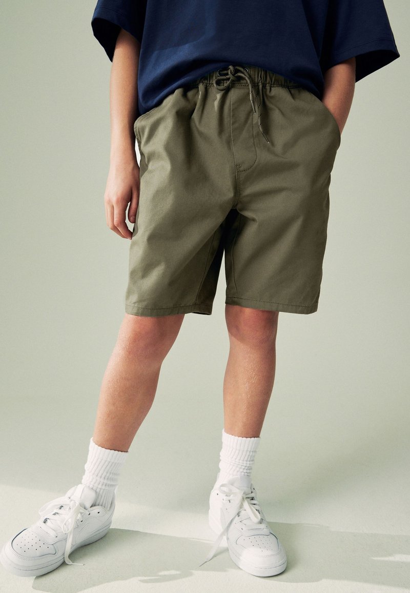 Shorts vert olive avec une taille élastique et un cordon de serrage. Associés à un t-shirt bleu marine ample, des baskets blanches et des chaussettes mi-mollet.