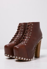 Botines de plataforma de cuero marrón con tacón grueso, con ojales de metal y tachuelas plateadas a lo largo de la suela. Cordones para un ajuste seguro.