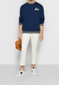 Sweatshirt bleu marine avec des accents jaunes, arborant deux motifs de sucettes glacées. Associé à un pantalon blanc retroussé et des baskets blanches. Tenant un chapeau à motif marron.