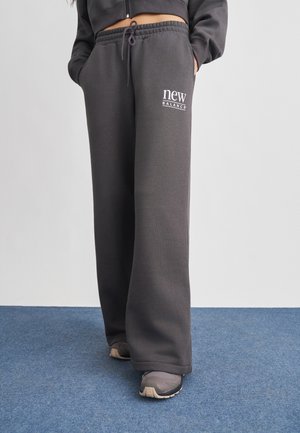 Osoba ubrana w ciemnoszare spodnie dresowe New Balance i trampki, stojąca na niebieskim dywanie, z rękami w kieszeniach.