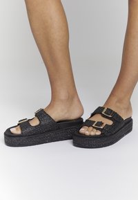 Sandali platform neri con tomaia testurizzata, due dettagli con fibbie e design open-toe. La robusta suola in gomma offre maggiore altezza e comfort.