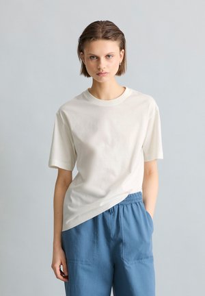 Jeune personne aux cheveux courts et bruns, portant un t-shirt blanc uni et un pantalon bleu ample, debout devant un fond gris clair.