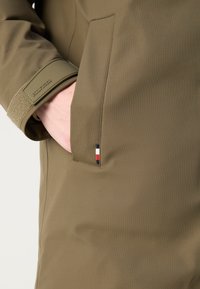 Olivgrüne, wasserdichte Jacke mit Strukturfinish, verstellbarem Bündchen, Seitentasche und einem kleinen Logodetail mit rot, weiß und blau Akzenten.