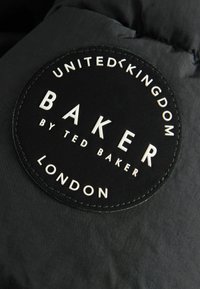 Écusson circulaire en tissu noir avec un texte blanc indiquant United Kingdom, Baker by Ted Baker, London sur un fond textile sombre.