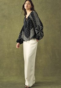 Blouse noire à motifs avec des manches volumineuses, présentant des motifs géométriques blancs. Associée à un pantalon large crème et des sandales à talons noires.