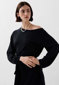 Černý top s dlouhým rukávem a designem bez ramínek, vyrobený z měkké látky. Má zavázaný pas a model, který nosí masivný stříbrný řetízkový náhrdelník.