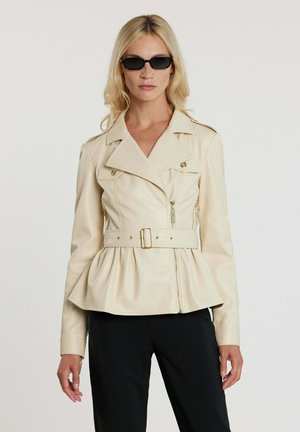 Donna che indossa giacca in pelle beige con cintura e baschina, pantaloni neri e piccoli occhiali da sole rettangolari neri, in piedi davanti a sfondo bianco.